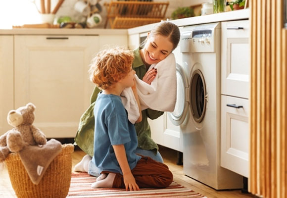 Cheerful family: kid son helping mother with linen in basket while doing laundry near the washing machine in the flight kitchen on the weekend at home